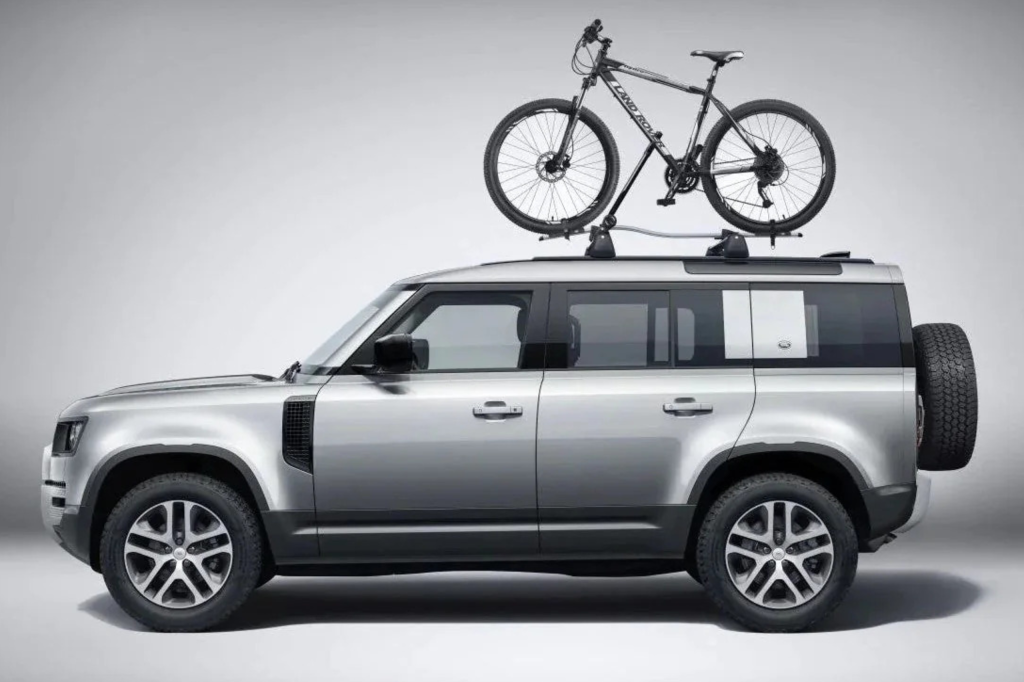 Perfil lateral de un Land Rover Defender plateado con una bicicleta de montaña negra montada en el portaequipajes del techo.