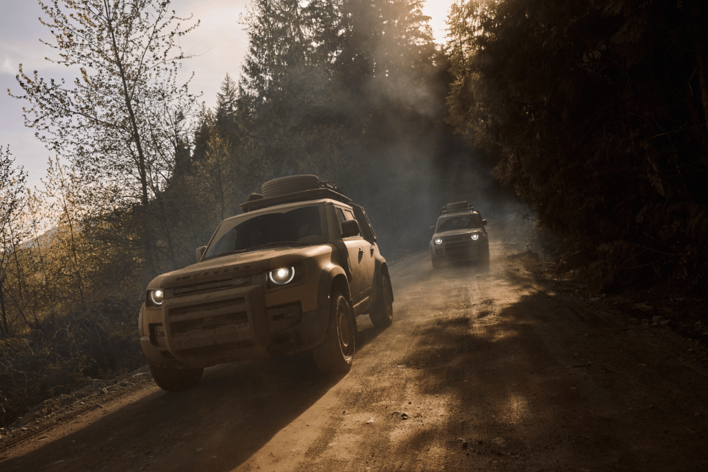 DEFENDER Trophy Edition en caravana off-road atravesando un camino de tierra entre bosque, con polvo y luz de atardecer