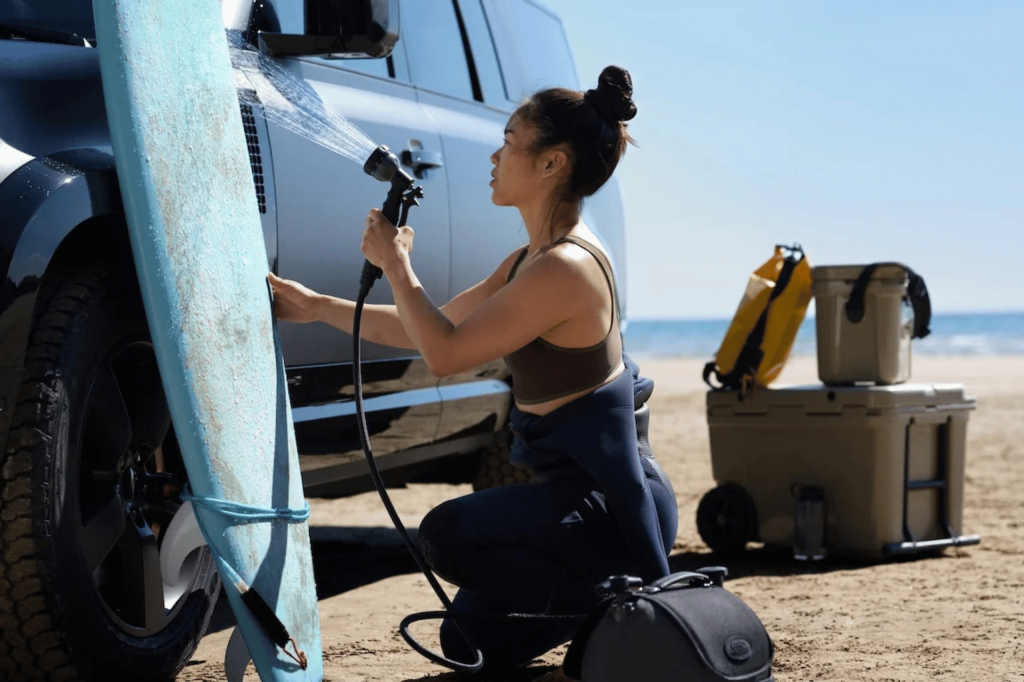 Mujer lavando su tabla de surf con el sistema de lavado portátil Land Rover junto a un DEFENDER en la playa